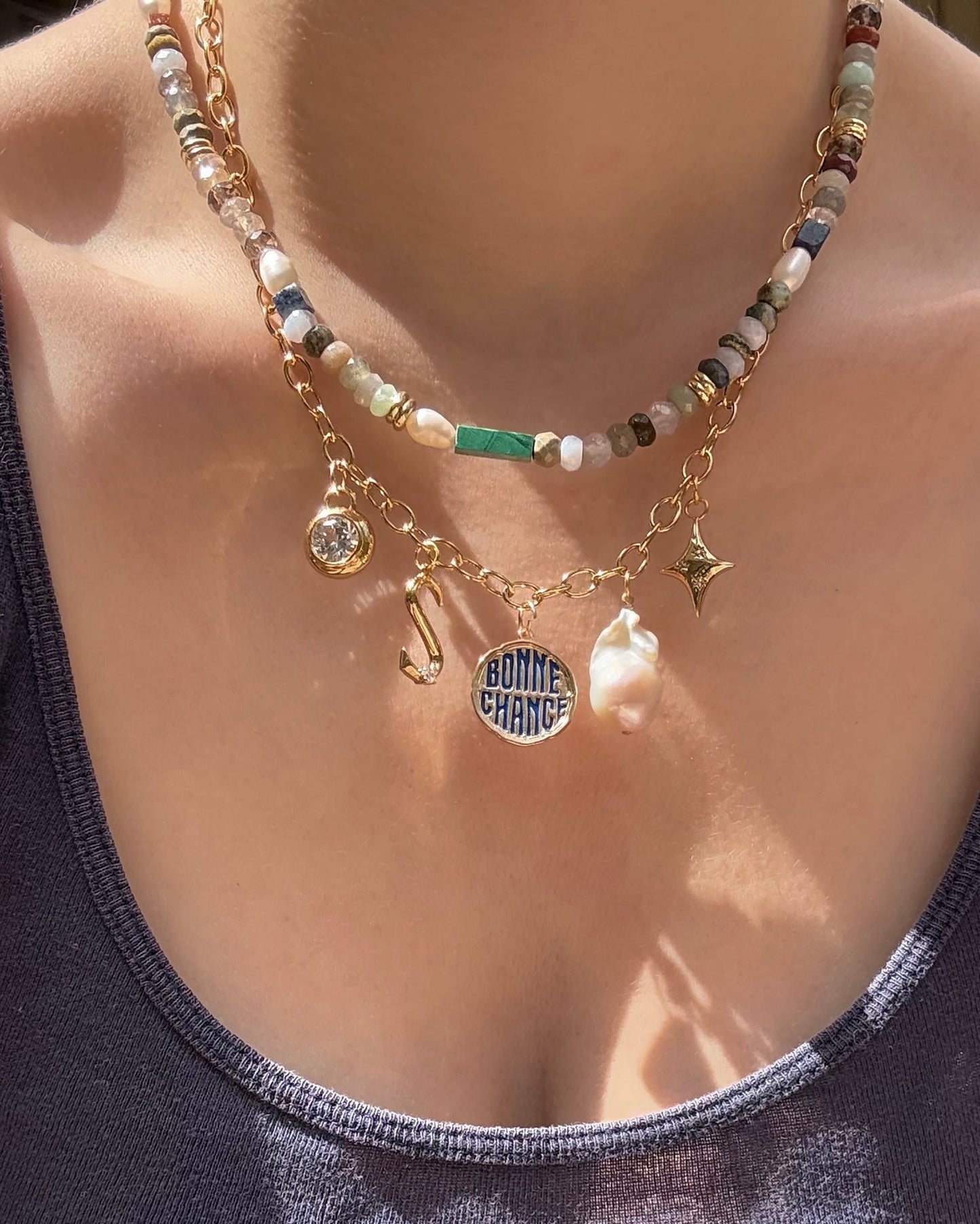 A close-up shows a person wearing layered necklaces—a beaded choker and a gold chain with charms including a BONNE CHANCE pendant, pearl, gemstone, cubic zirconia charm, hook, and the Mod + Jo Celeste Puffy Star Charm—over a dark tank top.