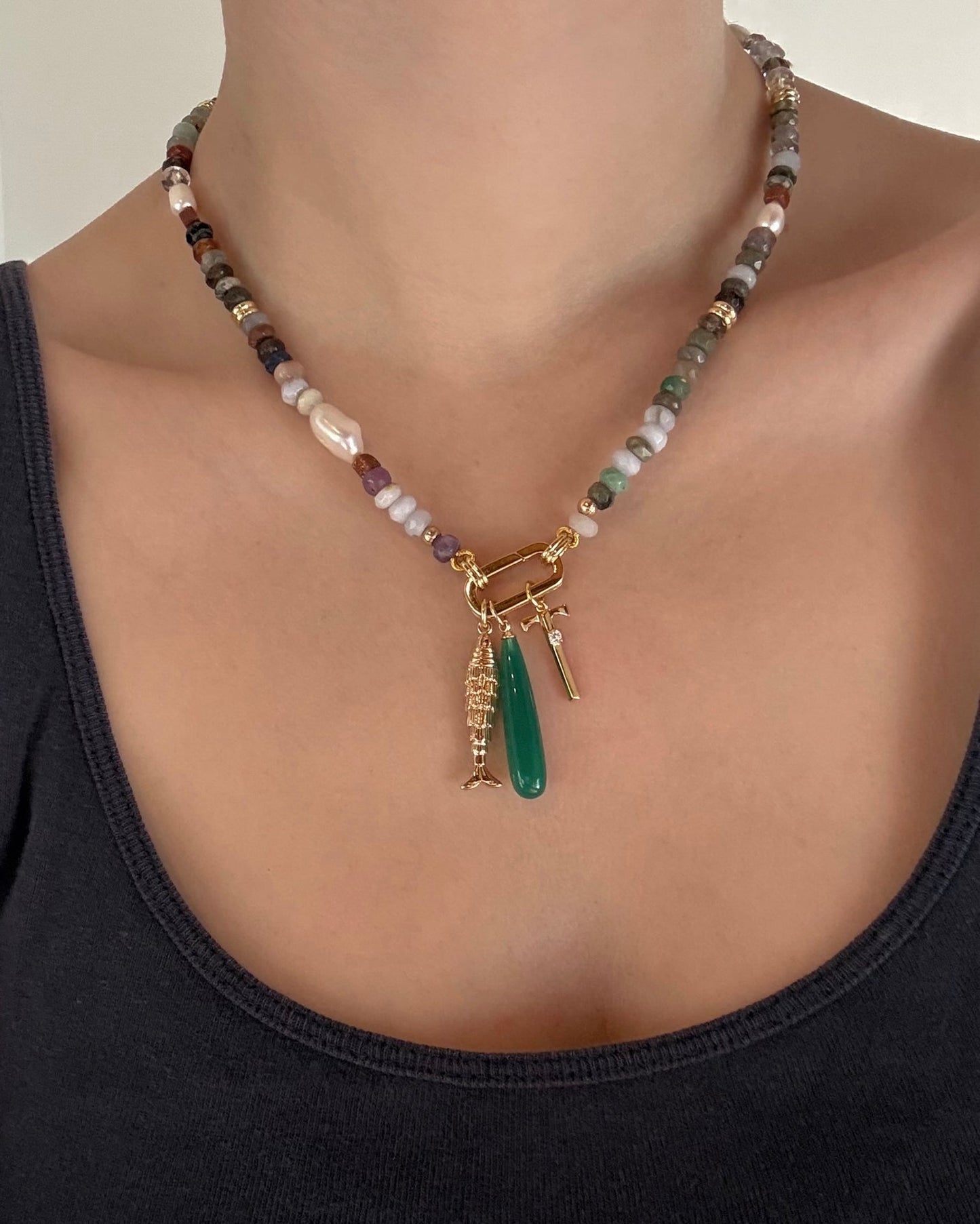 A person wears the Mod + Jo Eloise Letter Charm—a multicolored beaded necklace with gold-plated charms, including an initial and green pendant—paired with a dark tank top. Photo shows only the neck and upper chest.