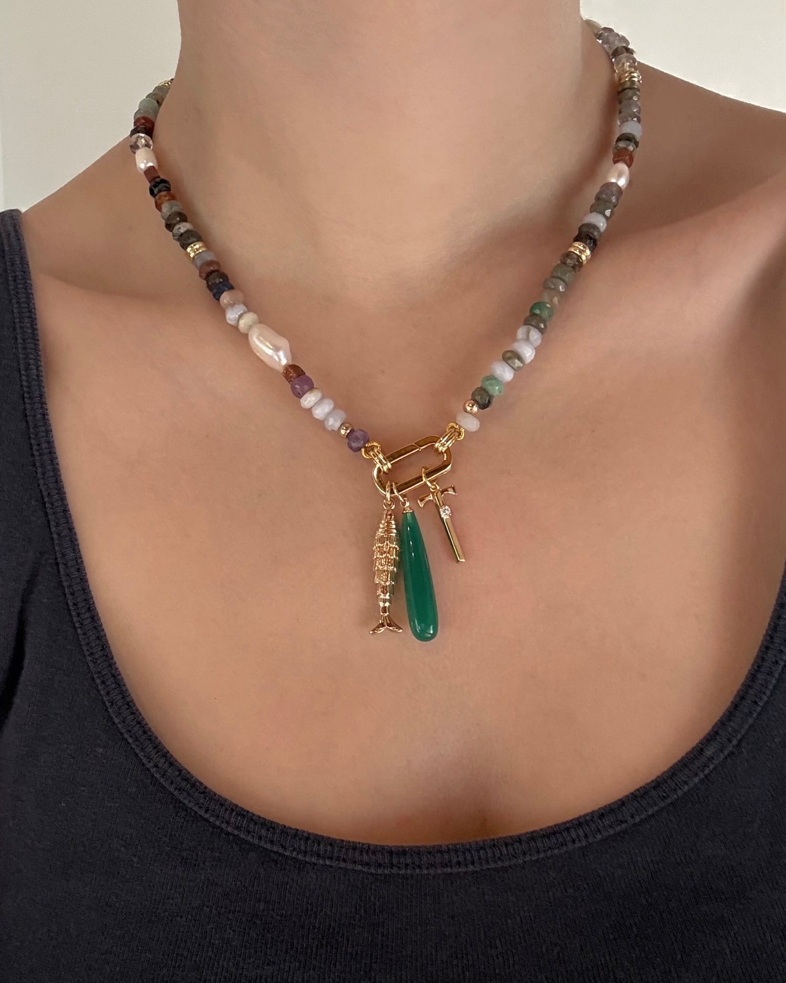 A person wears the Mod + Jo Eloise Letter Charm—a multicolored beaded necklace with gold-plated charms, including an initial and green pendant—paired with a dark tank top. Photo shows only the neck and upper chest.