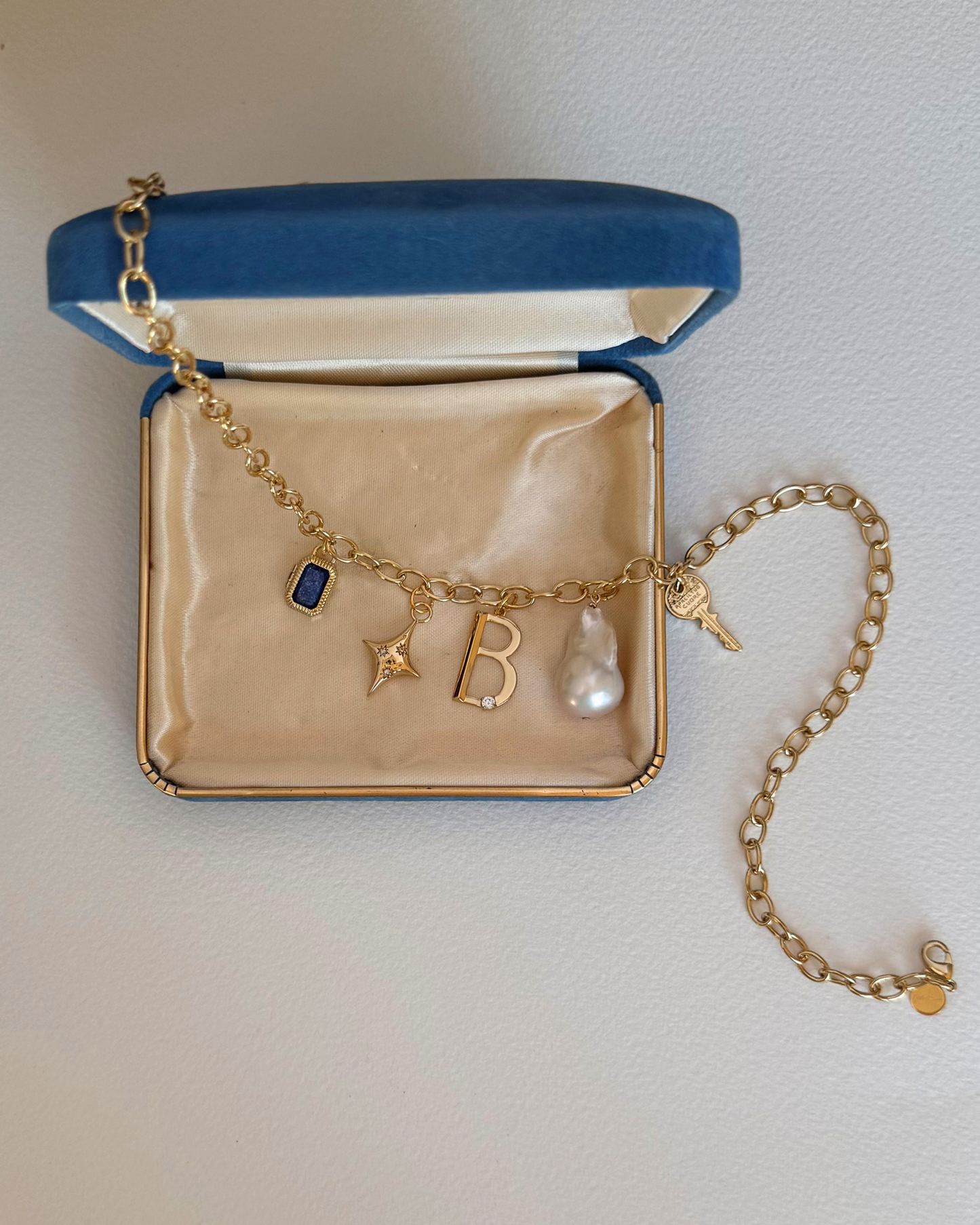 A Mod + Jo gold charm bracelet featuring the Celeste Puffy Star Charm, blue gem, letter "B," pearl, and key, displayed in an open blue velvet jewelry box with a cream-colored interior.