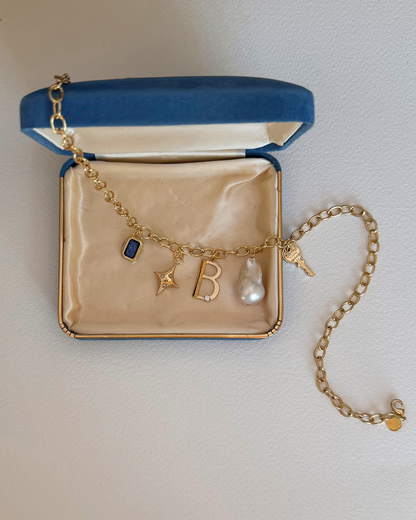 A Mod + Jo gold charm bracelet featuring the Celeste Puffy Star Charm, blue gem, letter "B," pearl, and key, displayed in an open blue velvet jewelry box with a cream-colored interior.