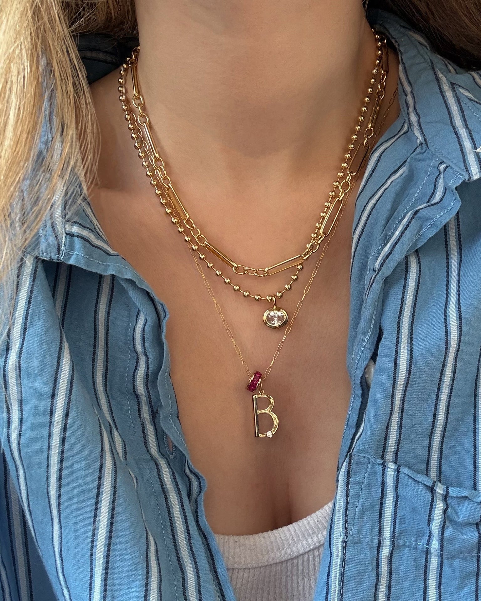 A woman wearing Mod + Jo's Soren Bezel Pendant Necklace—14k gold plated and nickel-free with a beaded chain, chunky link, and B initial pendant with a red stone—layered over a blue striped shirt and white top.