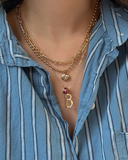 A person wears a blue striped shirt and three gold necklaces, including a gold plated letter B pendant with a red gem and the Mod + Jo Este Birthstone Charm Spacer.