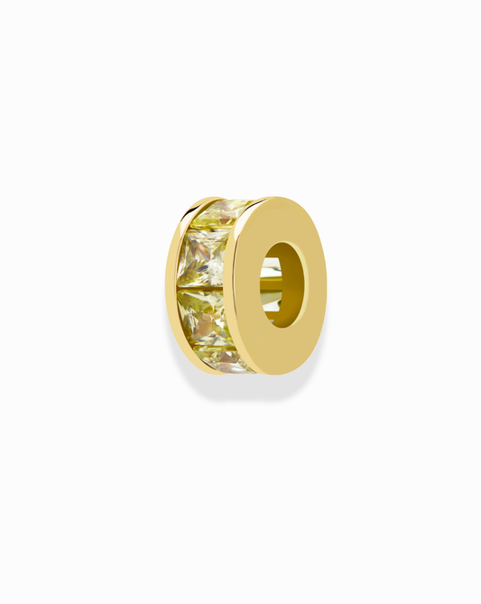 The Mod + Jo Este Birthstone Charm Spacer is a round gold bead with a hollow center and a band of yellow, princess-cut gemstones—ideal for personalized jewelry. Shown on a white background.