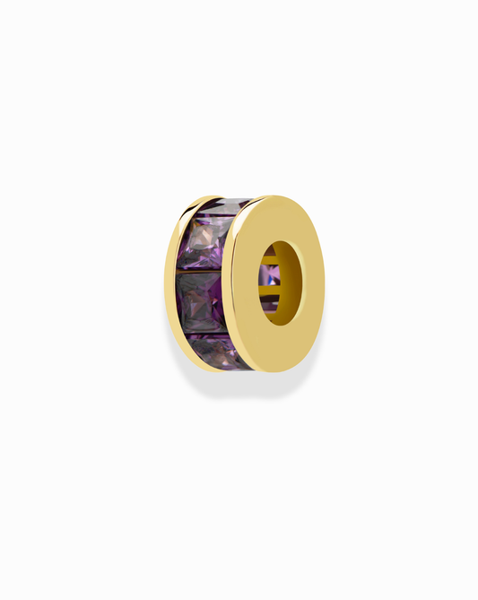 The Mod + Jo Este Birthstone Charm Spacer is a gold bead with a hollow center, featuring a row of rectangular purple gemstones around the edge—ideal for personalizing necklaces. Shown on a plain white background.
