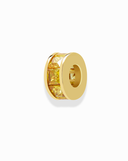 The Mod + Jo Este Birthstone Charm Spacer is a gold circular bead with a central row of square-cut yellow gemstones, ideal as a necklace spacer or birthstone charm. It’s shown photographed on a white background.