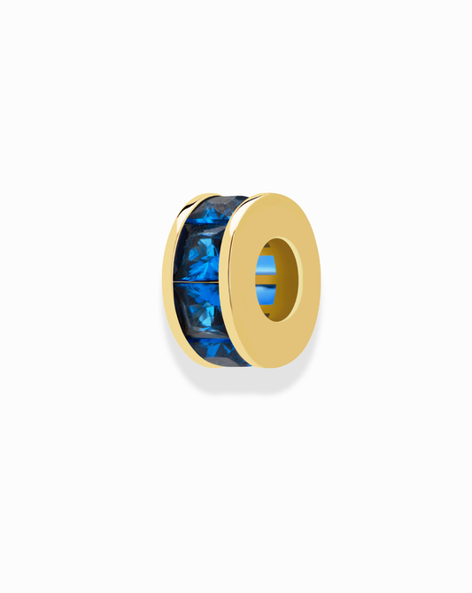 The Mod + Jo Este Birthstone Charm Spacer features a gold bead design with a faceted blue crystal band, ideal as a bracelet or necklace spacer, shown on a white background.