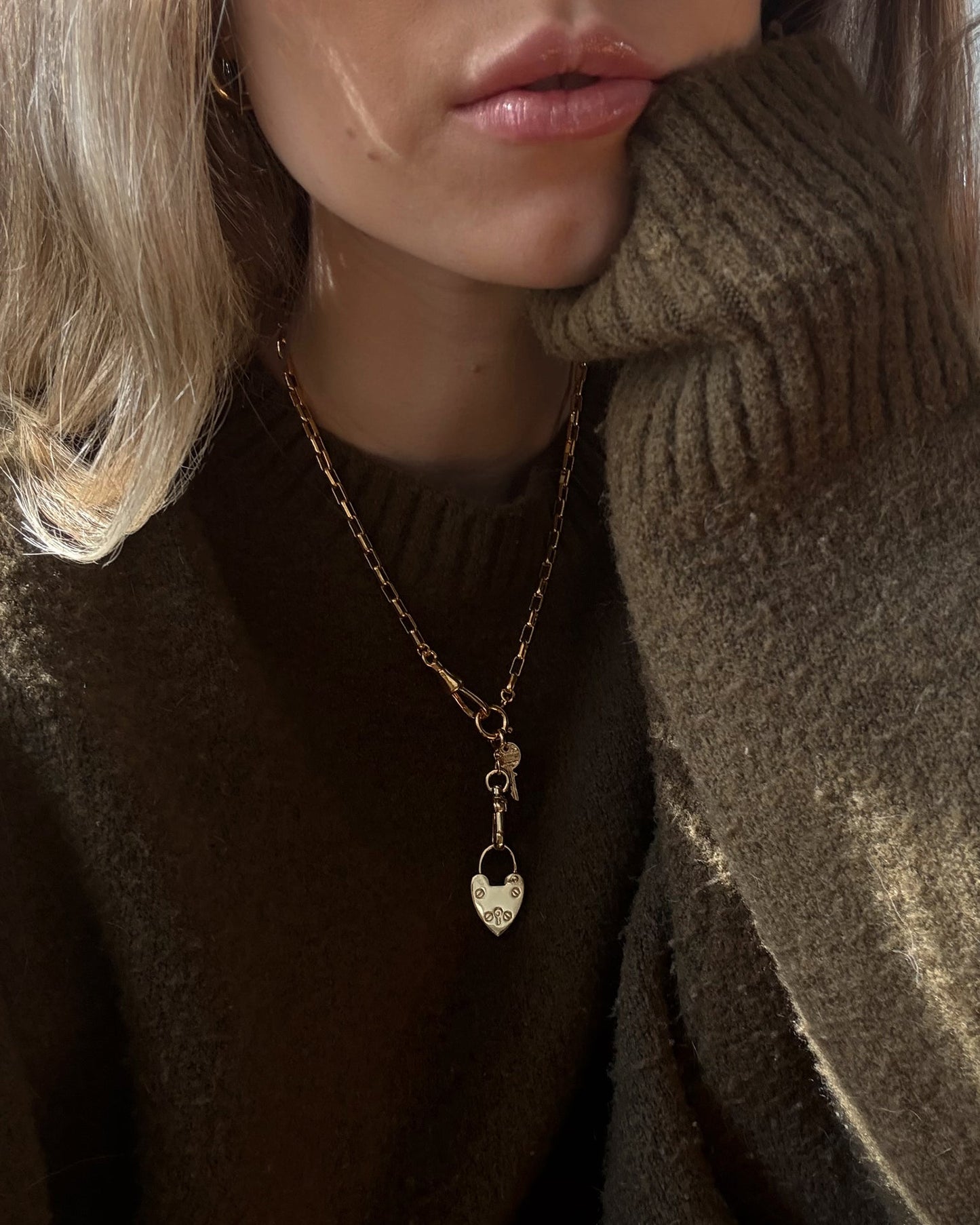 A woman in a chunky brown sweater rests her chin on her hand, showcasing the Mod + Jo Sasha Heart Locket Enhancer. Only her lower face and upper torso are visible.