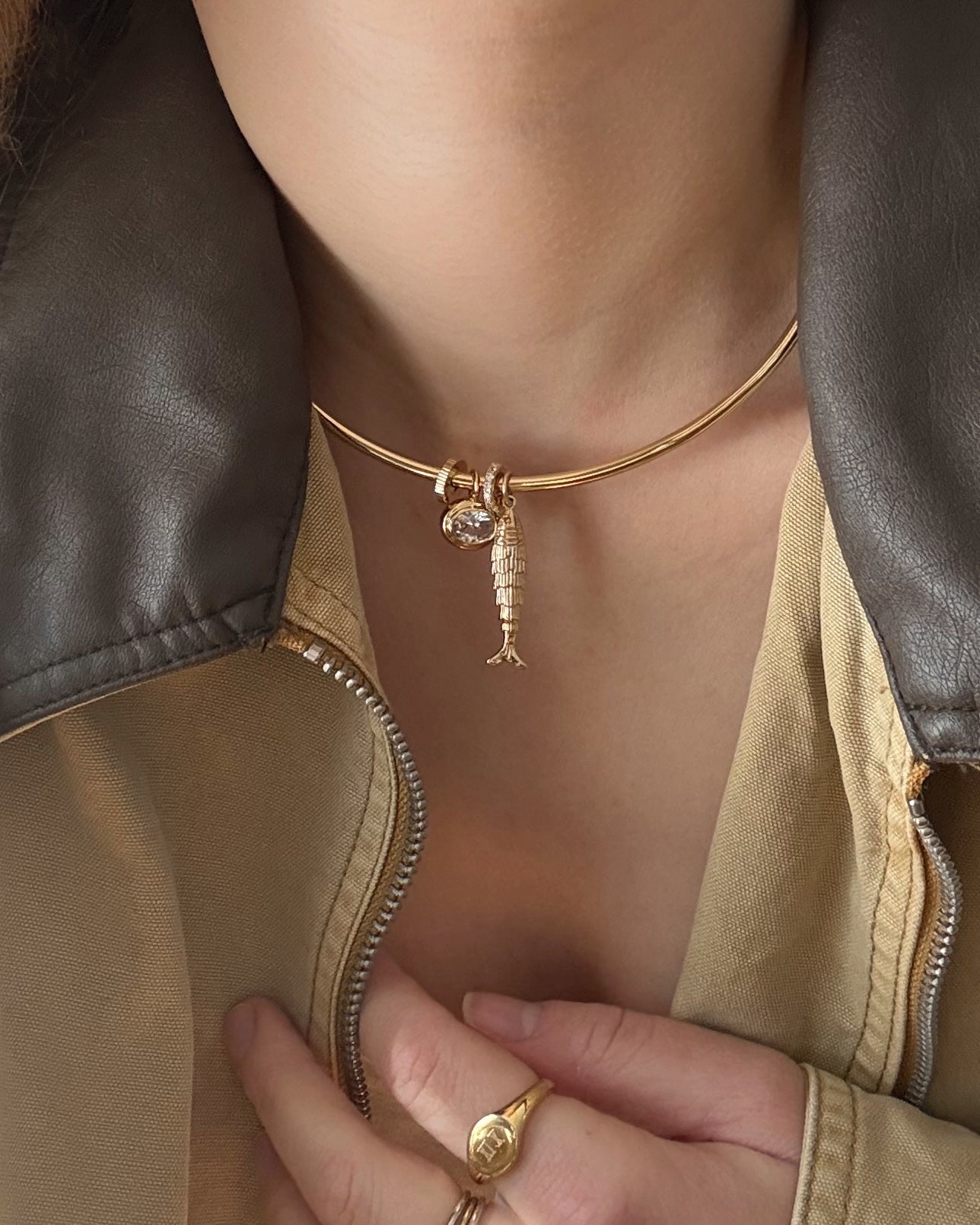 A person wears a Mod + Jo Shia Fish Charm 14k gold-plated choker with a round gem, paired with a beige jacket featuring a brown leather collar. Their hand, adorned with a gold ring, rests on their chest.