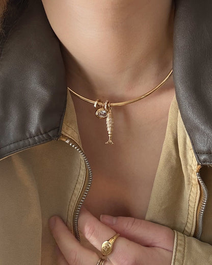 A person wears a Mod + Jo Shia Fish Charm 14k gold-plated choker with a round gem, paired with a beige jacket featuring a brown leather collar. Their hand, adorned with a gold ring, rests on their chest.