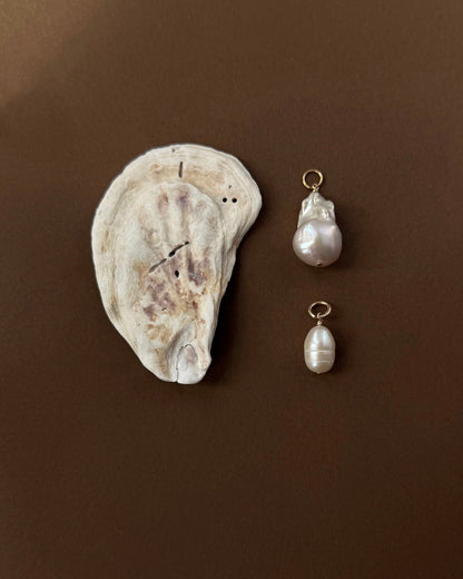 A flat, textured oyster shell rests on a brown surface beside two Mod + Jo charms: the Jaci Baroque Pearl Charm with an irregular baroque pearl and another featuring a smooth, oval freshwater pearl—both 14k gold plated.