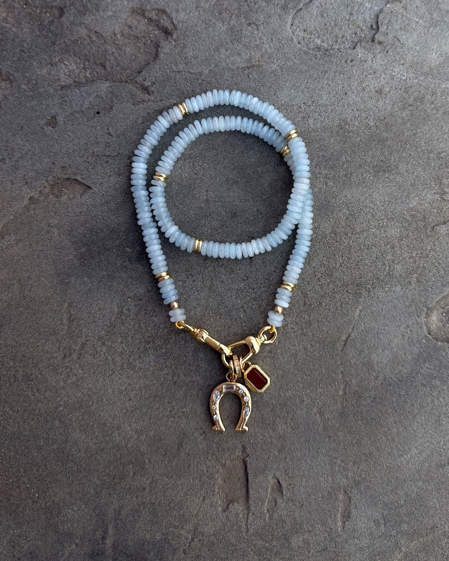The Sonia Gemstone Necklace by Mod + Jo features aquamarine beads, gold accents, and a rhinestone horseshoe charm—a stunning piece of handmade jewelry displayed on a textured gray surface.