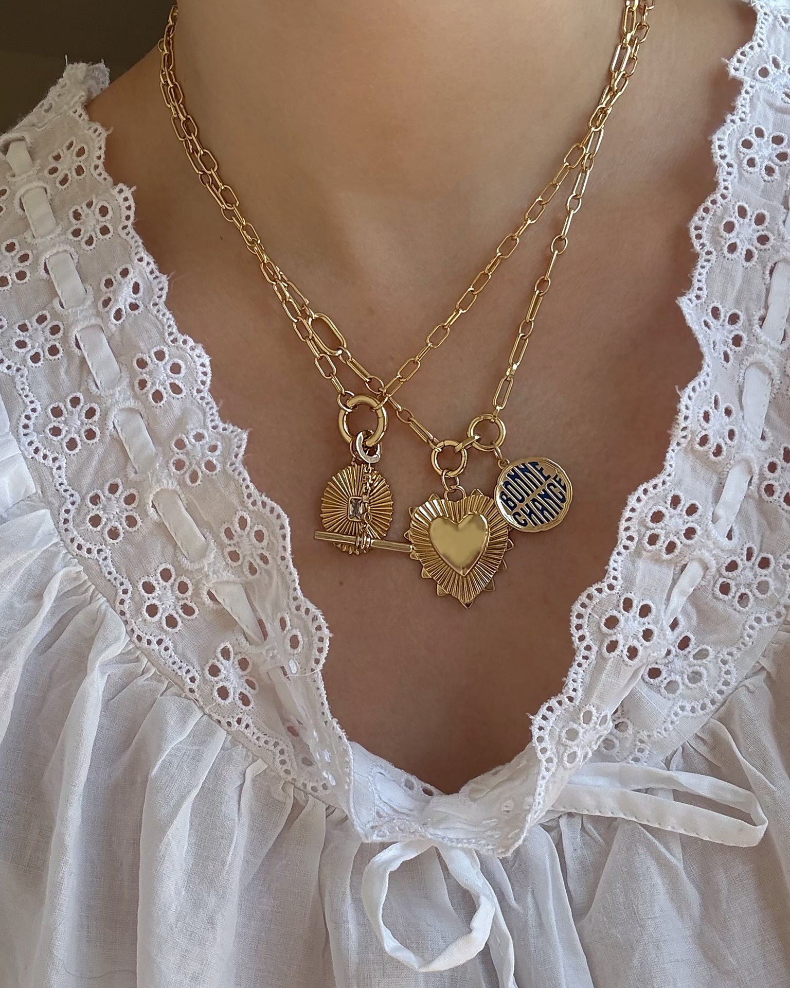 A person wearing a white eyelet blouse and two layered gold necklaces featuring three pendants: a sunburst, the Mod + Jo Grande Lynn Engravable Heart Charm in 14k gold over brass, and a round charm engraved with bonne chance.