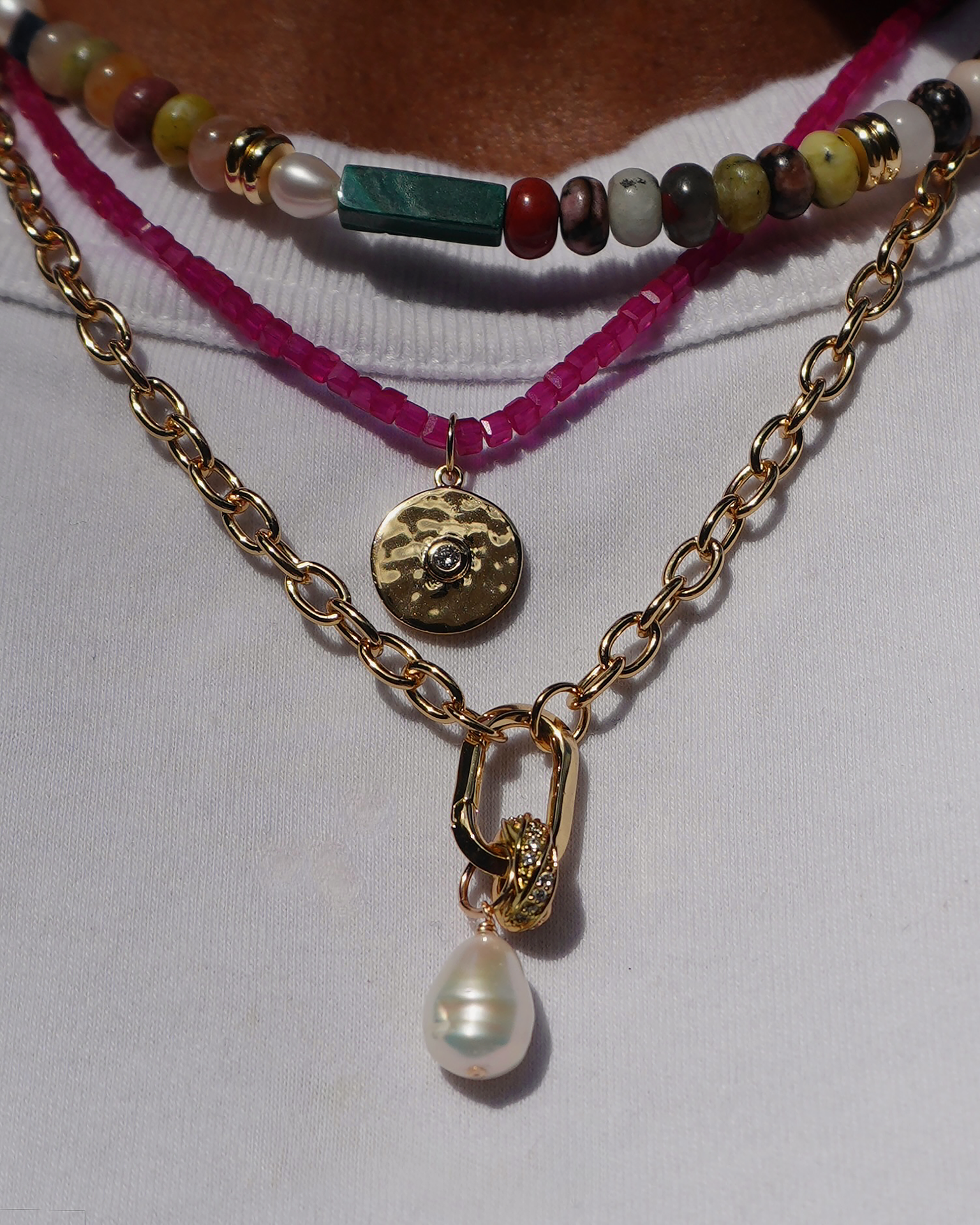 A close-up of layered necklaces: a colorful beaded choker, the Merit Gemstone Necklace with a round gold pendant, and a 14k gold-filled chain with a large pearl—all elegantly styled over a white shirt.