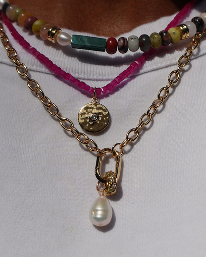 A close-up of layered necklaces: a colorful beaded choker, the Merit Gemstone Necklace with a round gold pendant, and a 14k gold-filled chain with a large pearl—all elegantly styled over a white shirt.