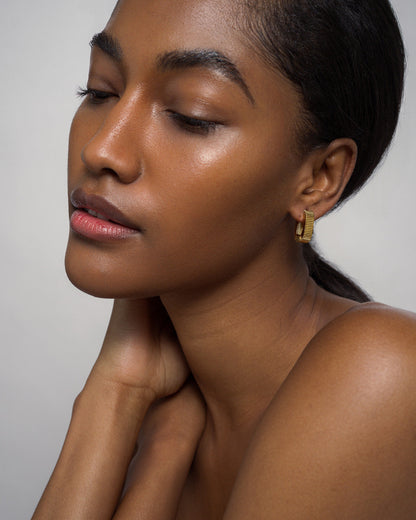 A woman with smooth, glowing skin poses with her eyes closed, hand gently touching her neck. She wears the Elly Fluted Hoops by Mod + Jo—lightweight gold earrings that complement her pulled-back dark hair. The background is plain and light-colored.