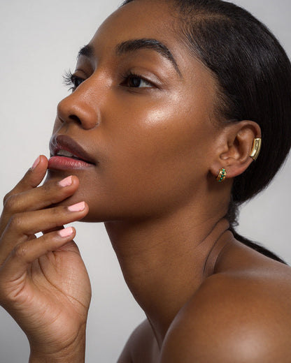 A woman with glowing skin and natural makeup poses in profile, gently touching her face. She wears Mod + Jo's Evy Raised Gemstone Hoops with 18k gold plating and a gold ear cuff. Her nails are soft pink against a neutral background.