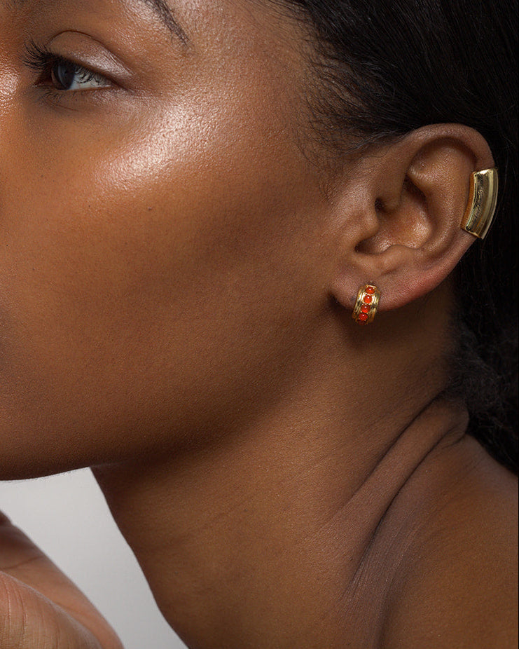 Close-up of a person with smooth brown skin and short hair wearing Mod + Jo’s Evy Raised Gemstone Hoops, featuring small gold hoops with red accents and a larger 18k gold-plated ear cuff.