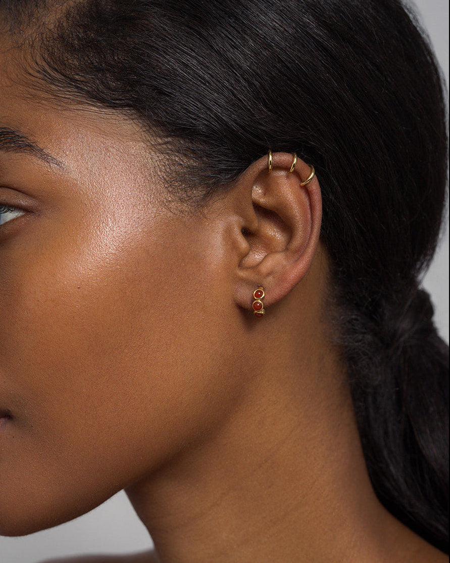 Close-up of a woman's left ear wearing Mod + Jo's Corynn Gemstone Huggies—three 18k gold plated hoops and a small stud with natural red agate. Her dark hair is pulled back, highlighting her smooth, radiant skin.