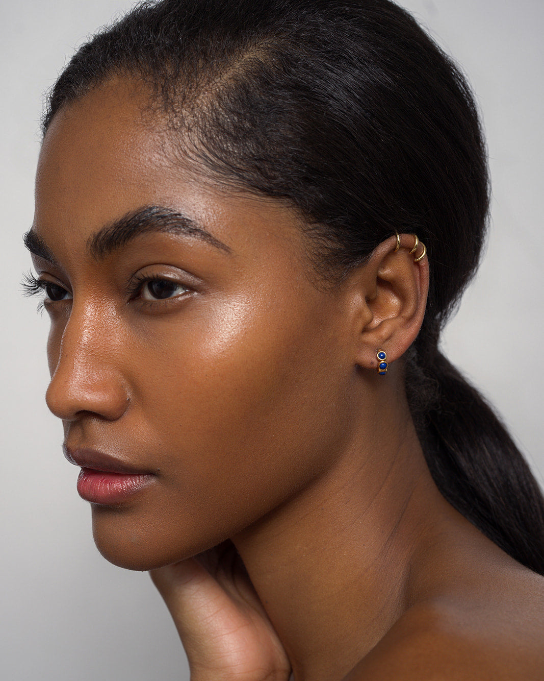 A woman with smooth, glowing skin and dark hair pulled back wears Mod + Jo's Corynn Gemstone Huggies—18k gold plated with natural gemstones—while gazing thoughtfully to the side against a neutral background.