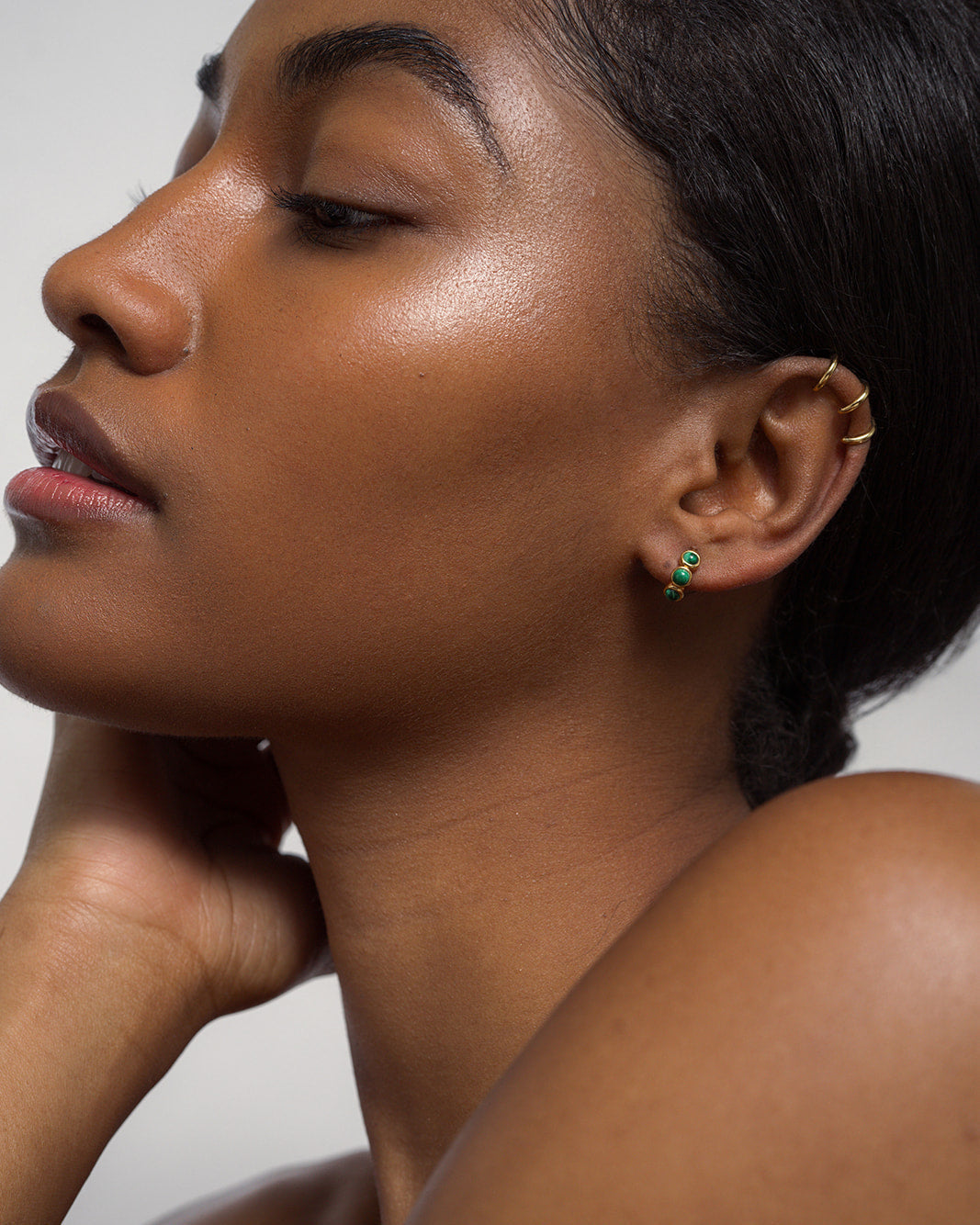 A close-up of a woman with glowing skin, her hair pulled back, wearing Mod + Jo's Corynn Gemstone Huggies in 18k gold plate and matching ear cuffs, her hand resting gently on her neck.
