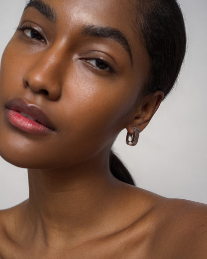 A woman with smooth brown skin and dark hair pulled back gazes softly at the camera. She wears a single, chunky Elly Fluted Hoop by Mod + Jo, paired with natural makeup and bare shoulders against a plain background. The bold silver earring adds a touch of modern elegance to her understated look.