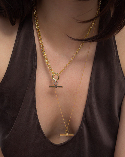 A person wearing a dark, deep V-neck top showcases two layered Mod + Jo Elliot Toggle Charm necklaces in 14k gold, each featuring a horizontal bar pendant and distinct chain styles, set against their bare upper chest.