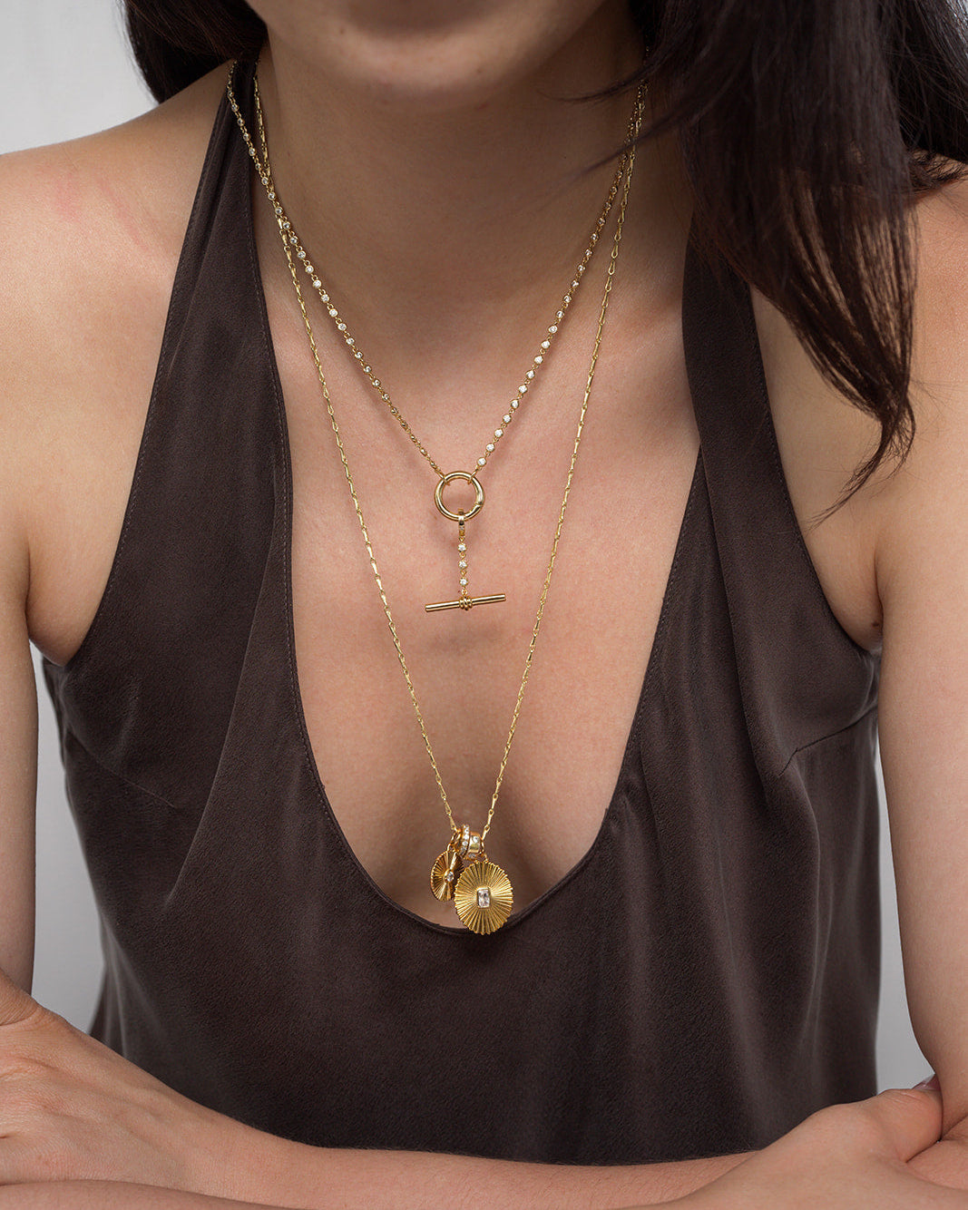 A woman wearing a deep brown V-neck top and layered Mod + Jo gold-plated jewelry, featuring the Livia Tennis Toggle Charm along with various pendants, including a CZ charm, a bar-accented circle, and disk- and sunburst hand-shaped charms.