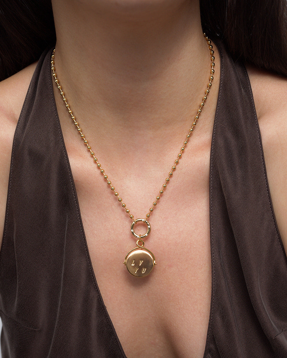 A person wearing a brown satin top and the Mod + Jo Bella "I Love You" Spinner Charm—a 14k gold necklace featuring a round pendant engraved with “LYU”—on a ball chain resting at the center of the chest.