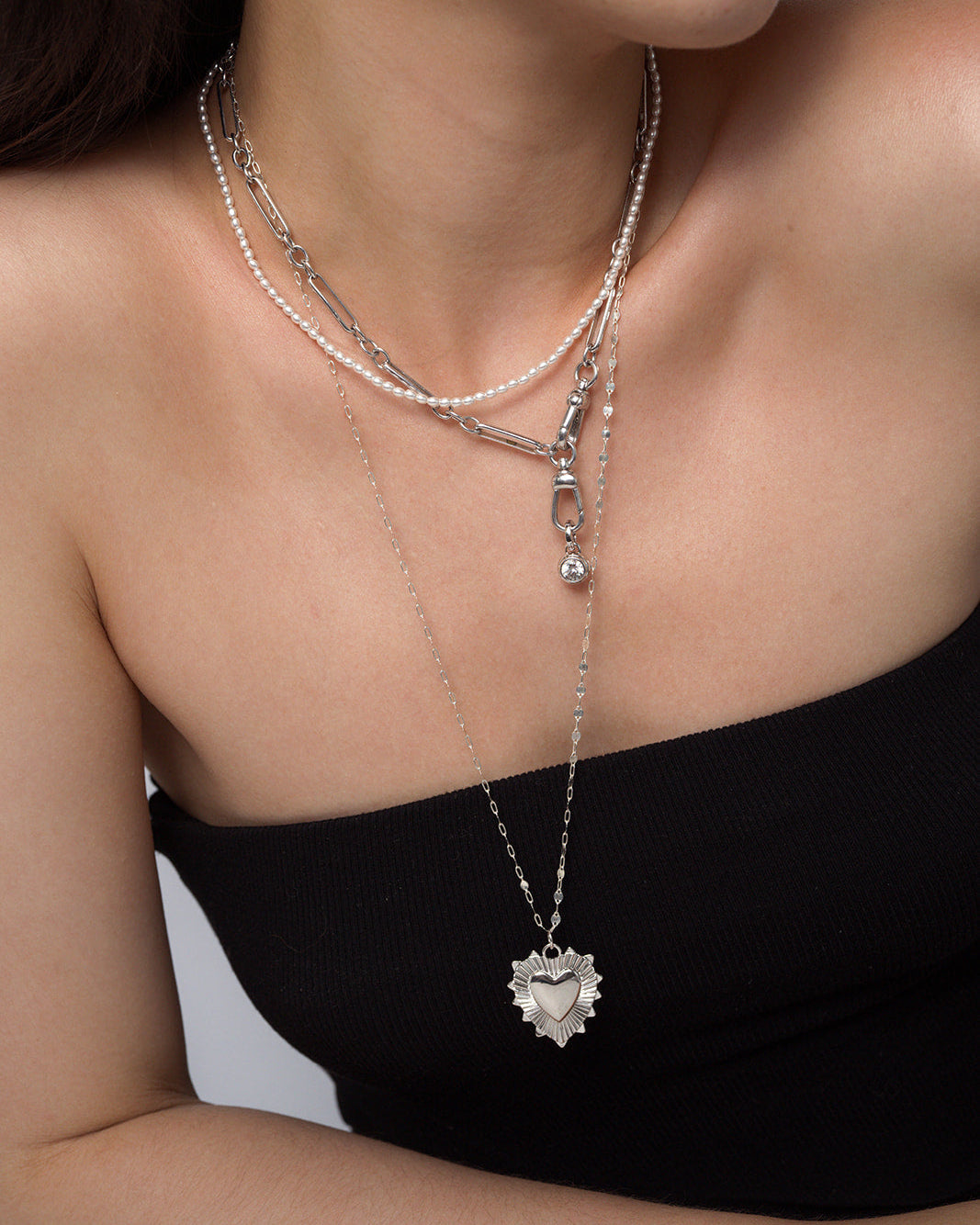 A woman wears a black off-shoulder top and layered silver necklaces, featuring the Mod + Jo Grande Lynn Engravable Heart Charm that adds a personalized touch to her elegant look.