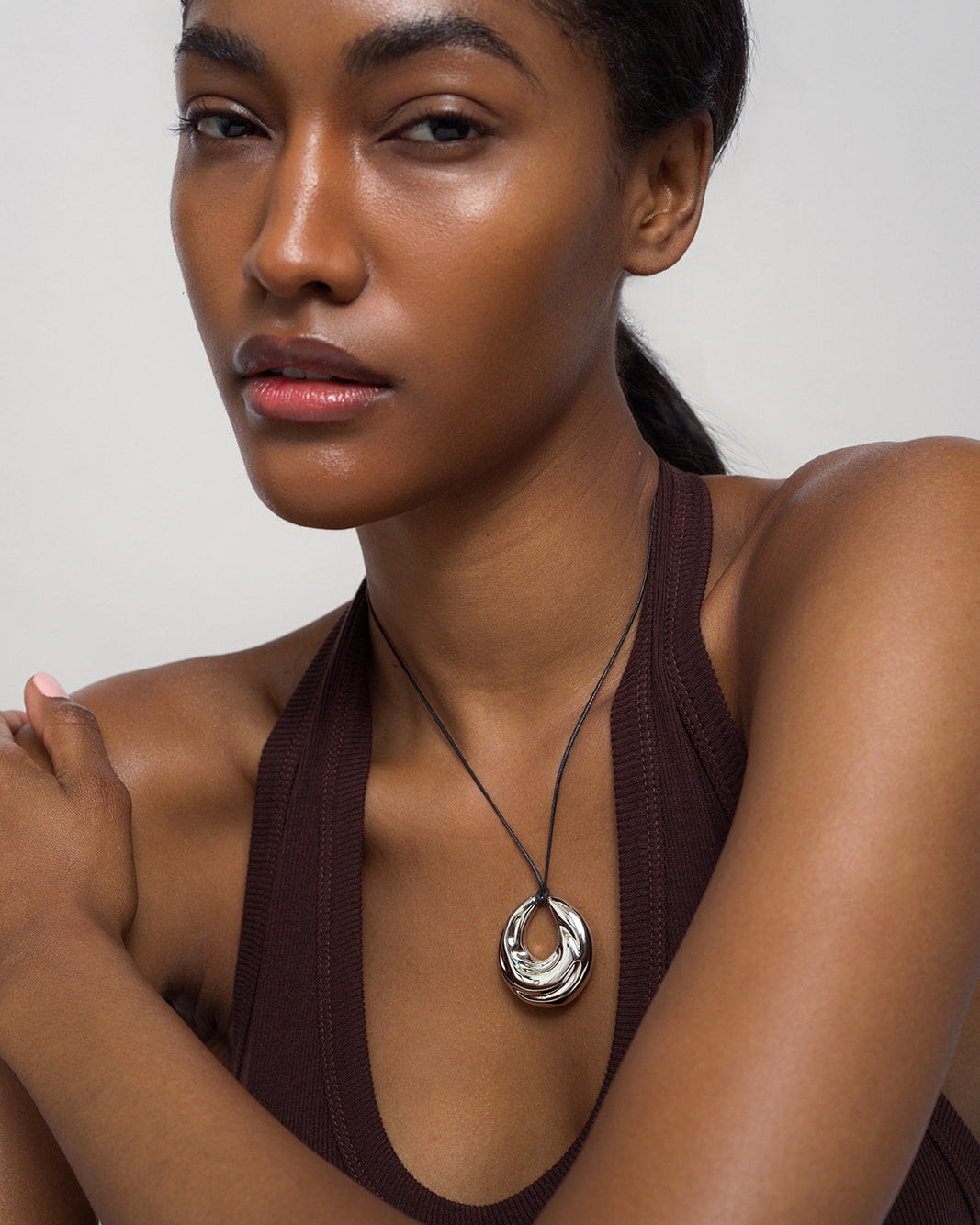 A woman in a sleeveless brown top poses with folded arms, showcasing the Mod + Jo Kira Cord Necklace—an adjustable gold and silver pendant style—against a plain background.