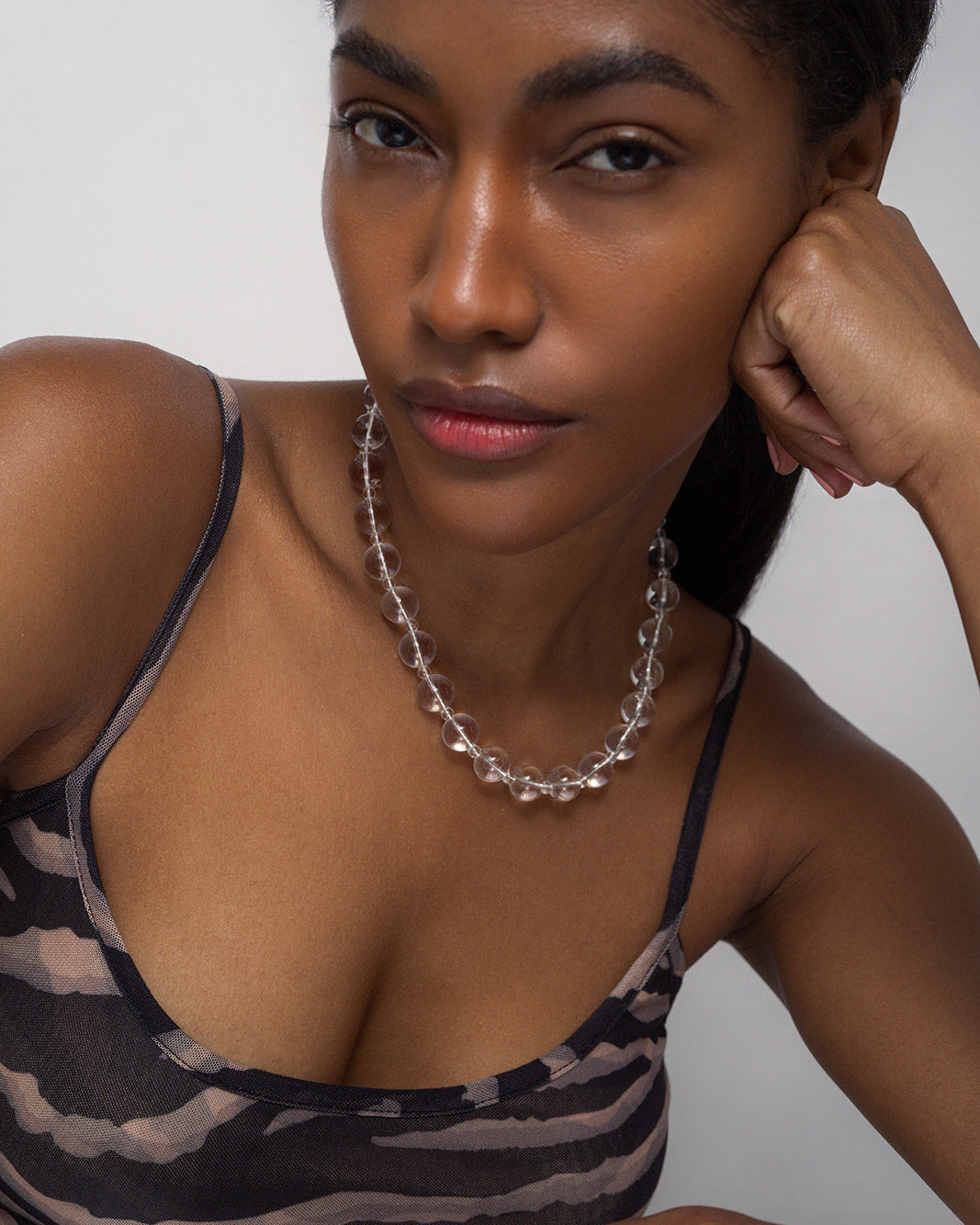 A woman with medium skin tone wears the Portia Gemstone Necklace by Mod + Jo and a sheer zebra-print tank top, posing confidently with her chin on her hand against a plain background.