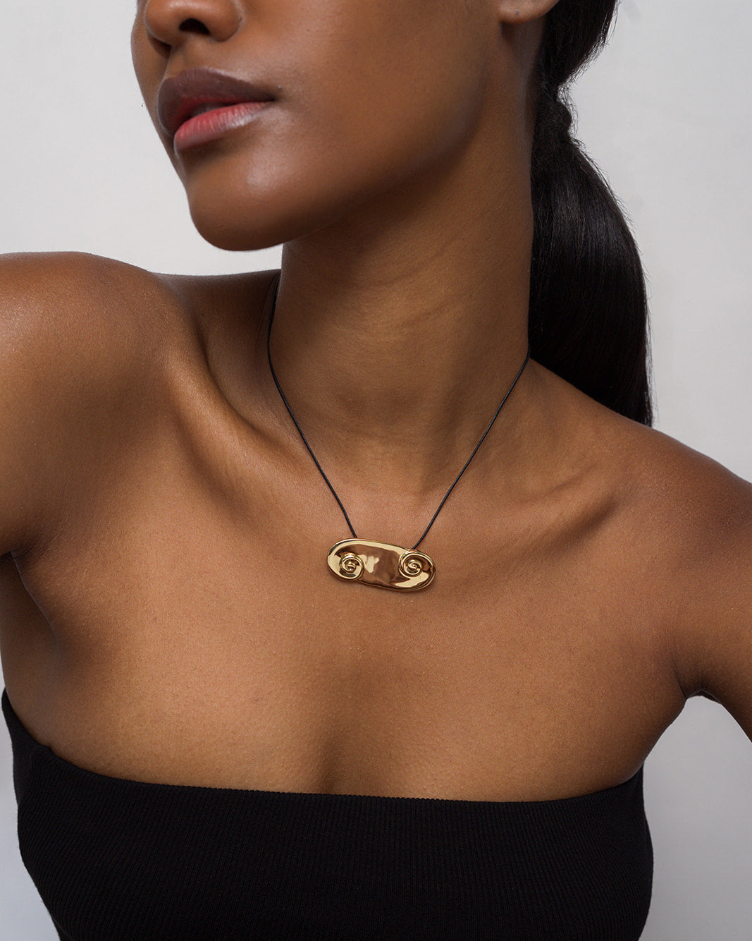 A woman wears the Mod + Jo *Preorder* Suna Spiral Cord Necklace, a 14k gold pendant with a unique abstract design on an adjustable bolo cord. She has sleek dark hair tied back and is dressed in a black strapless top.