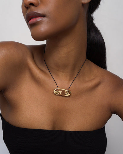 A woman wears the Mod + Jo *Preorder* Suna Spiral Cord Necklace, a 14k gold pendant with a unique abstract design on an adjustable bolo cord. She has sleek dark hair tied back and is dressed in a black strapless top.
