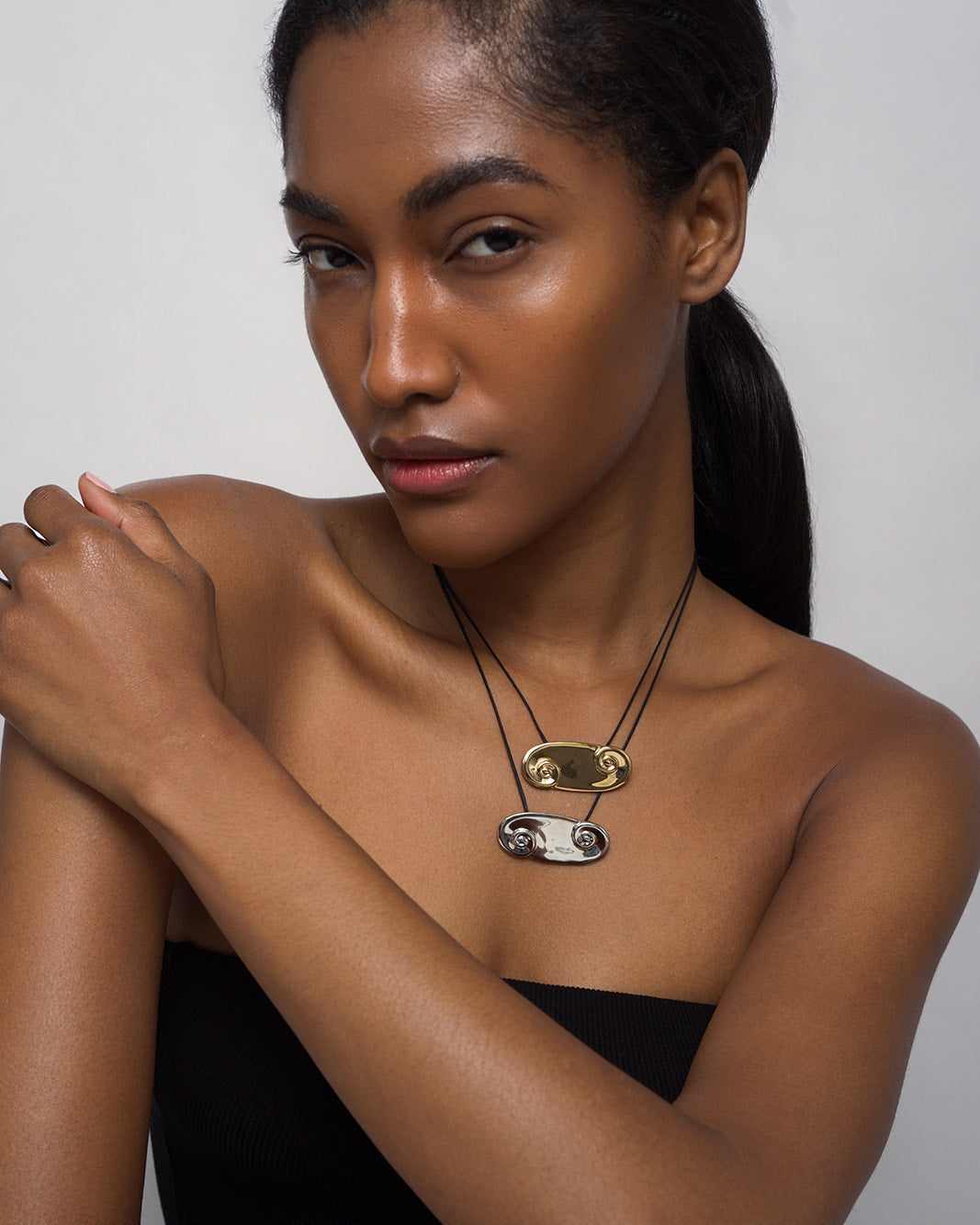 A woman with medium-dark skin and long, dark hair in a low ponytail poses against a light background, wearing a black strapless top and two Mod + Jo Suna Spiral Cord Necklaces—one with a 14k gold pendant—on adjustable bolo cords.