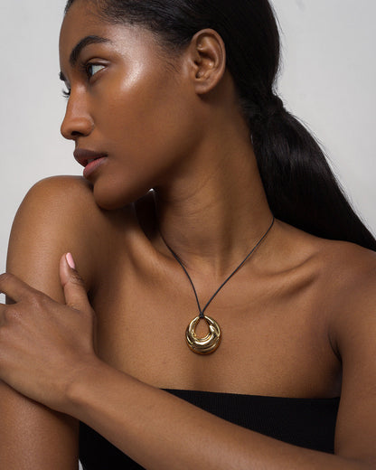 A woman with dark skin and long, dark hair in a low ponytail poses in profile, wearing a black strapless top and the Mod + Jo Kira Cord Necklace, featuring an adjustable cord and gold pendant, her arm gently resting across her chest.