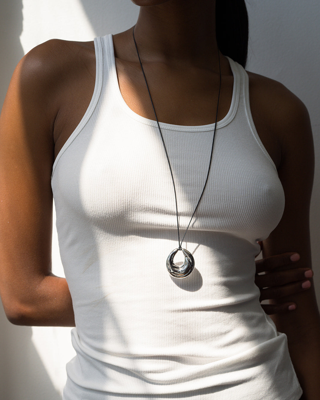 Wearing a white ribbed tank top, a person stands in partial sunlight with one hand on their side, showcasing the Mod + Jo Kira Cord Necklace featuring a large adjustable gold and silver pendant.