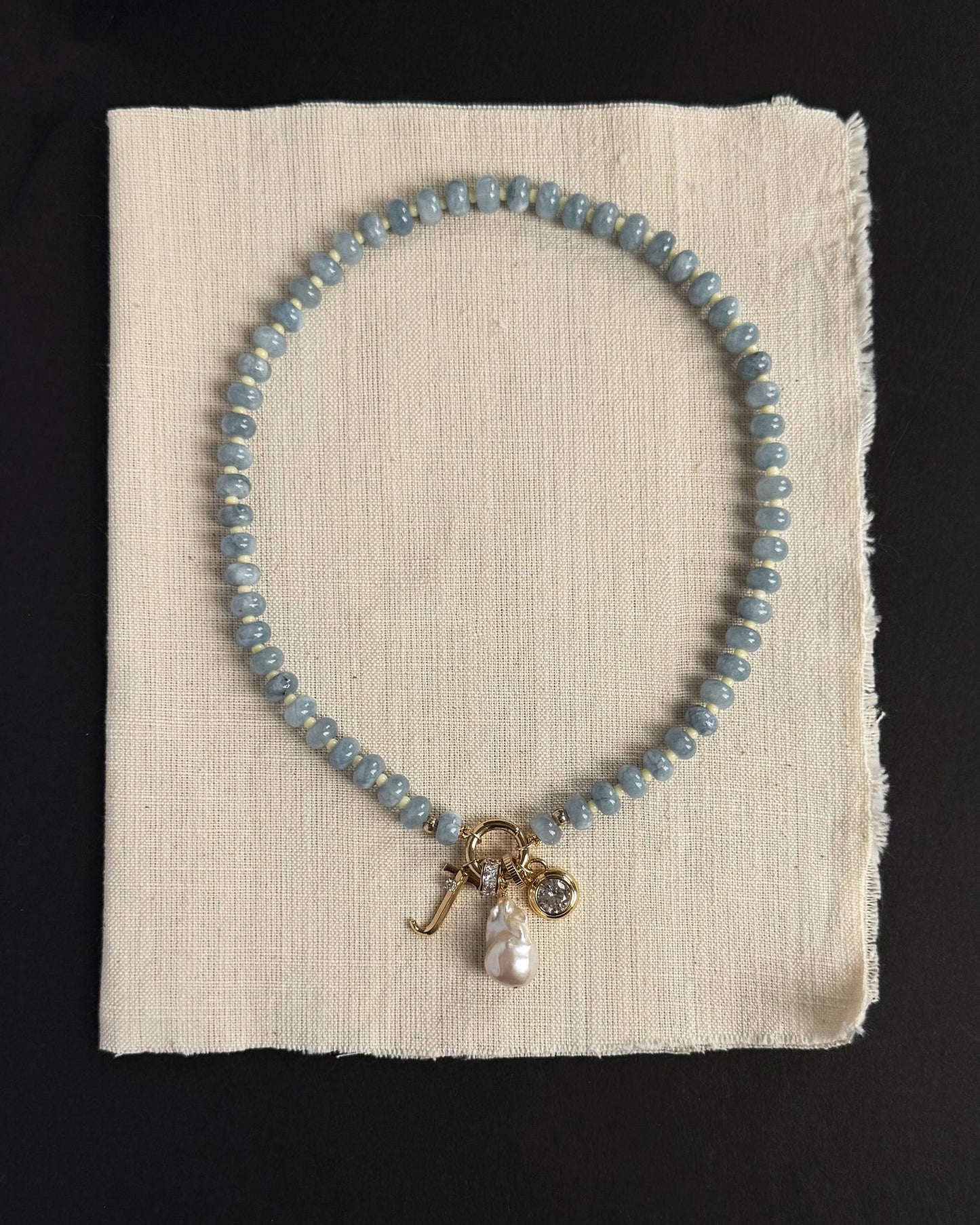 The Macey Gemstone Necklace by Mod + Jo, featuring blue beads, gold charms, a pearl, and a crystal pendant, is displayed on off-white fabric against a black background, highlighting the elegant appeal of genuine gemstones.