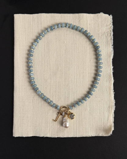 The Macey Gemstone Necklace by Mod + Jo, featuring blue beads, gold charms, a pearl, and a crystal pendant, is displayed on off-white fabric against a black background, highlighting the elegant appeal of genuine gemstones.