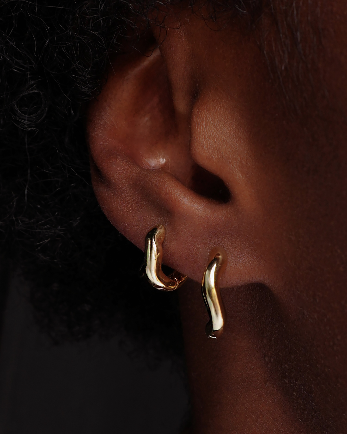 Close-up of person's ear with gold wavy huggies earrings. Jewelry product detail.