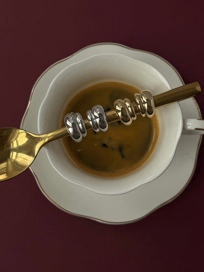 A cup of coffee on a white saucer with a gold spoon displays five Mod + Jo Petite Sofie Volume Hoops, their shiny metallic finish stacked elegantly on the spoon, set against a dark burgundy background.