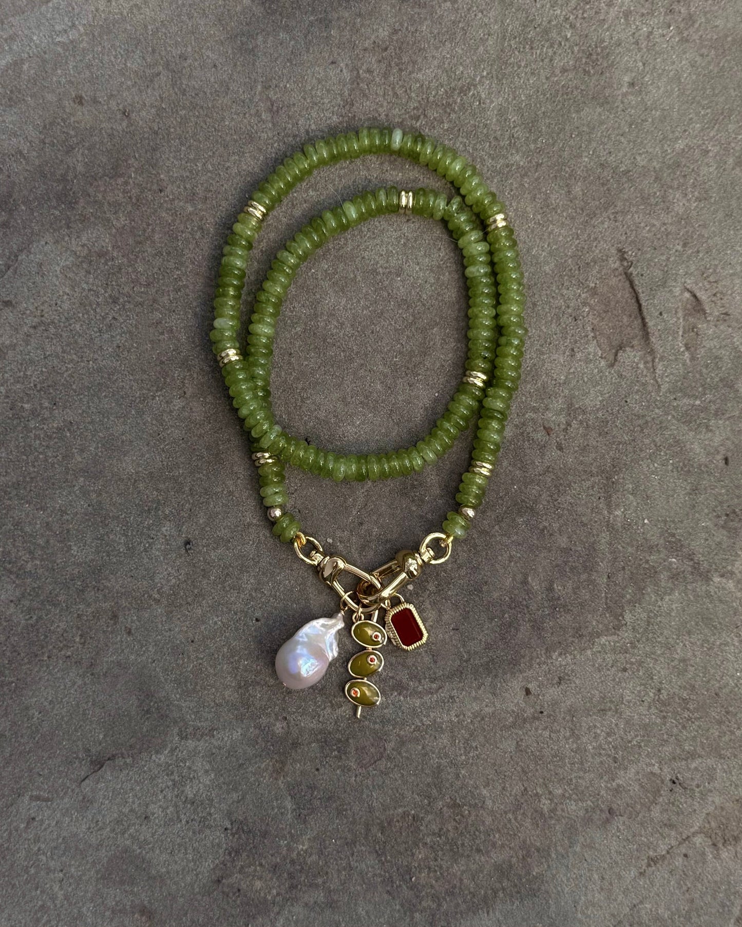 The Sonia Gemstone Necklace by Mod + Jo, crafted with genuine gemstone beads and gold accents, is displayed on a gray background. It features three stunning charms: a pearl, a gold spiral, and a red gem—ideal for handmade jewelry lovers.