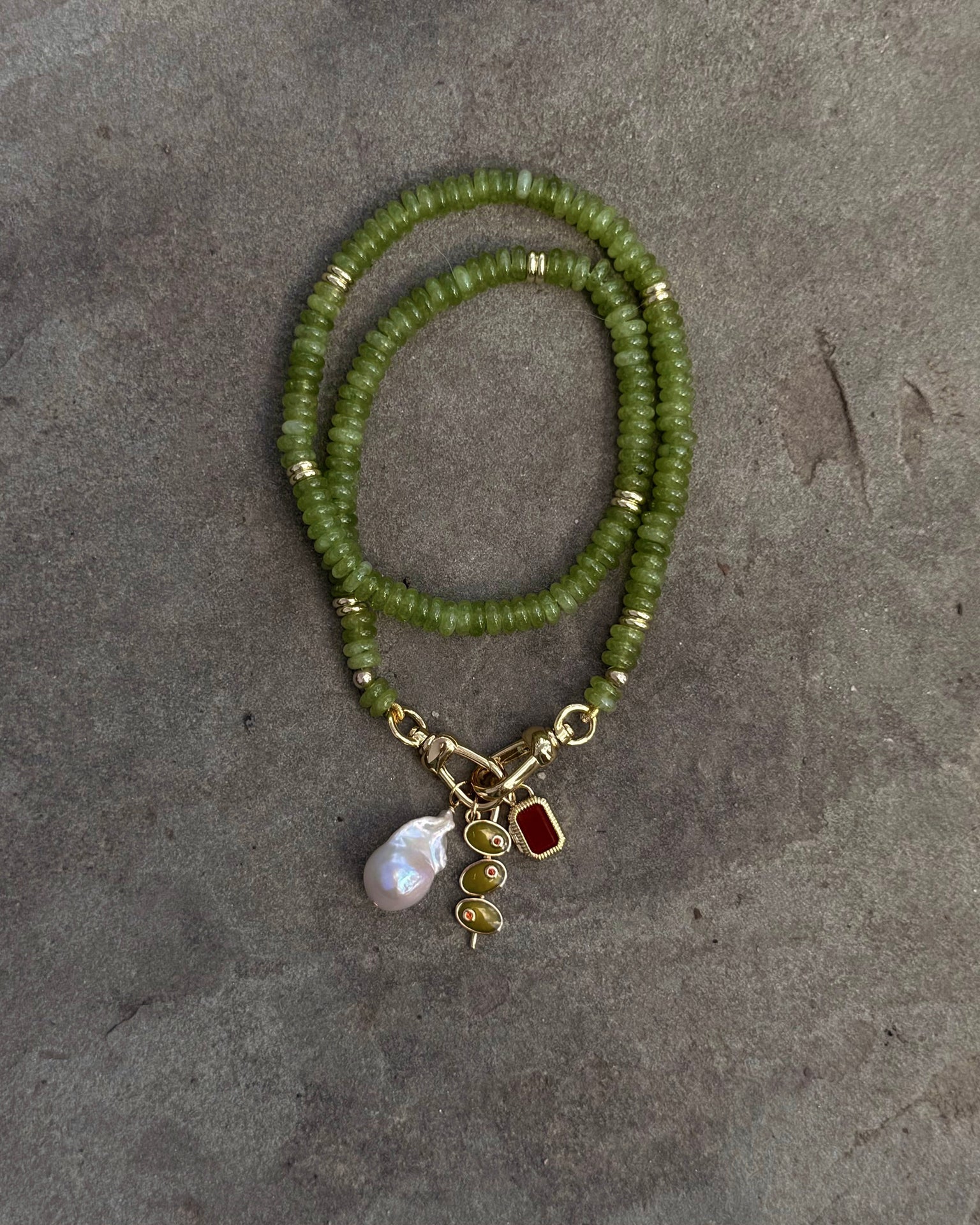 The Sonia Gemstone Necklace by Mod + Jo, crafted with genuine gemstone beads and gold accents, is displayed on a gray background. It features three stunning charms: a pearl, a gold spiral, and a red gem—ideal for handmade jewelry lovers.