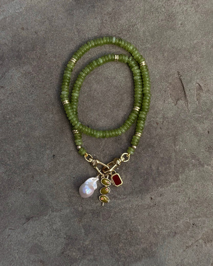 The Sonia Gemstone Necklace by Mod + Jo, crafted with genuine gemstone beads and gold accents, is displayed on a gray background. It features three stunning charms: a pearl, a gold spiral, and a red gem—ideal for handmade jewelry lovers.