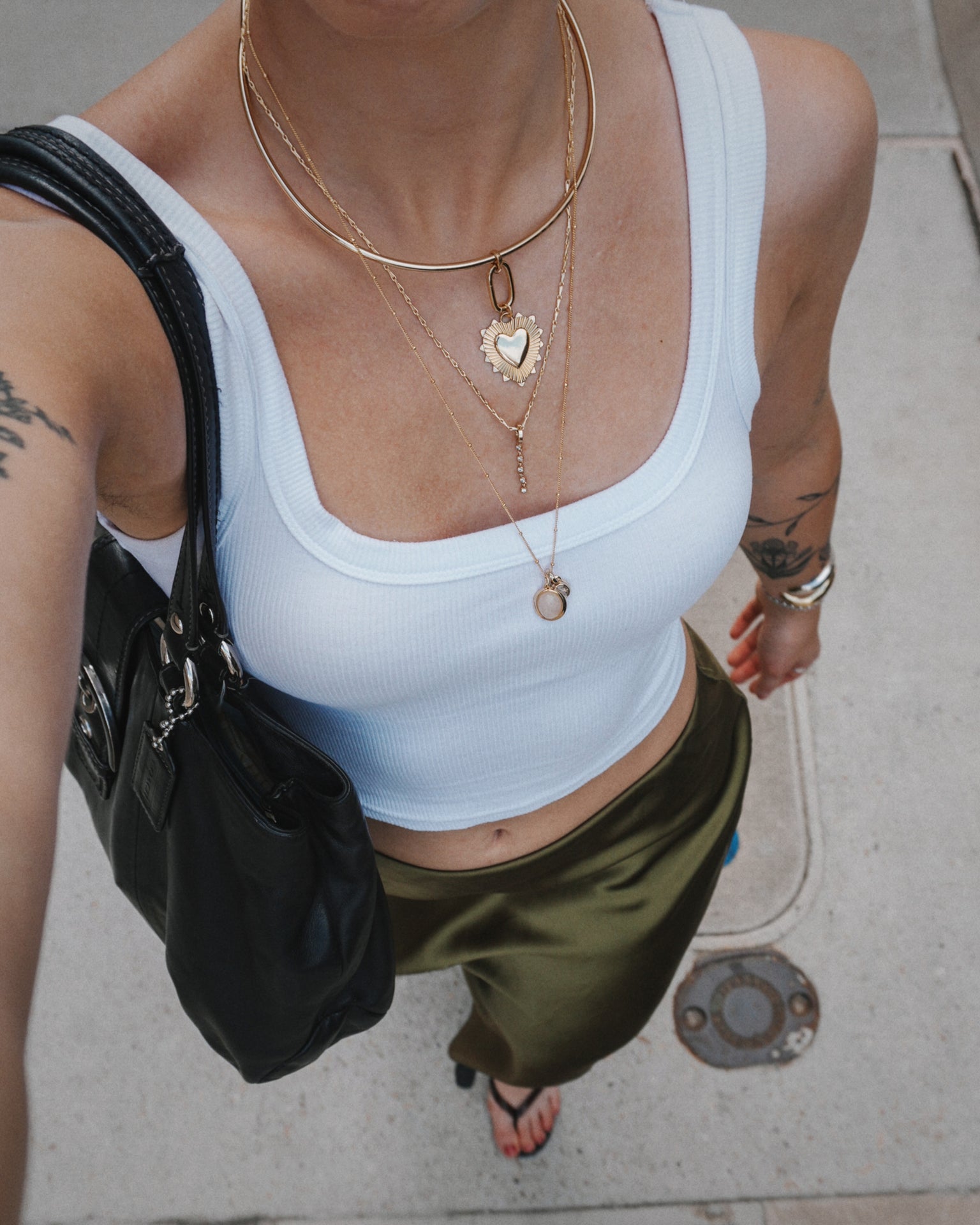 A person in a white tank top, olive green skirt, and black bag stands on a city sidewalk, wearing gold layered necklaces including the Mod + Jo Grande Lynn Engravable Heart Charm. Tattoos are visible on their arm. Photo is taken from above.
