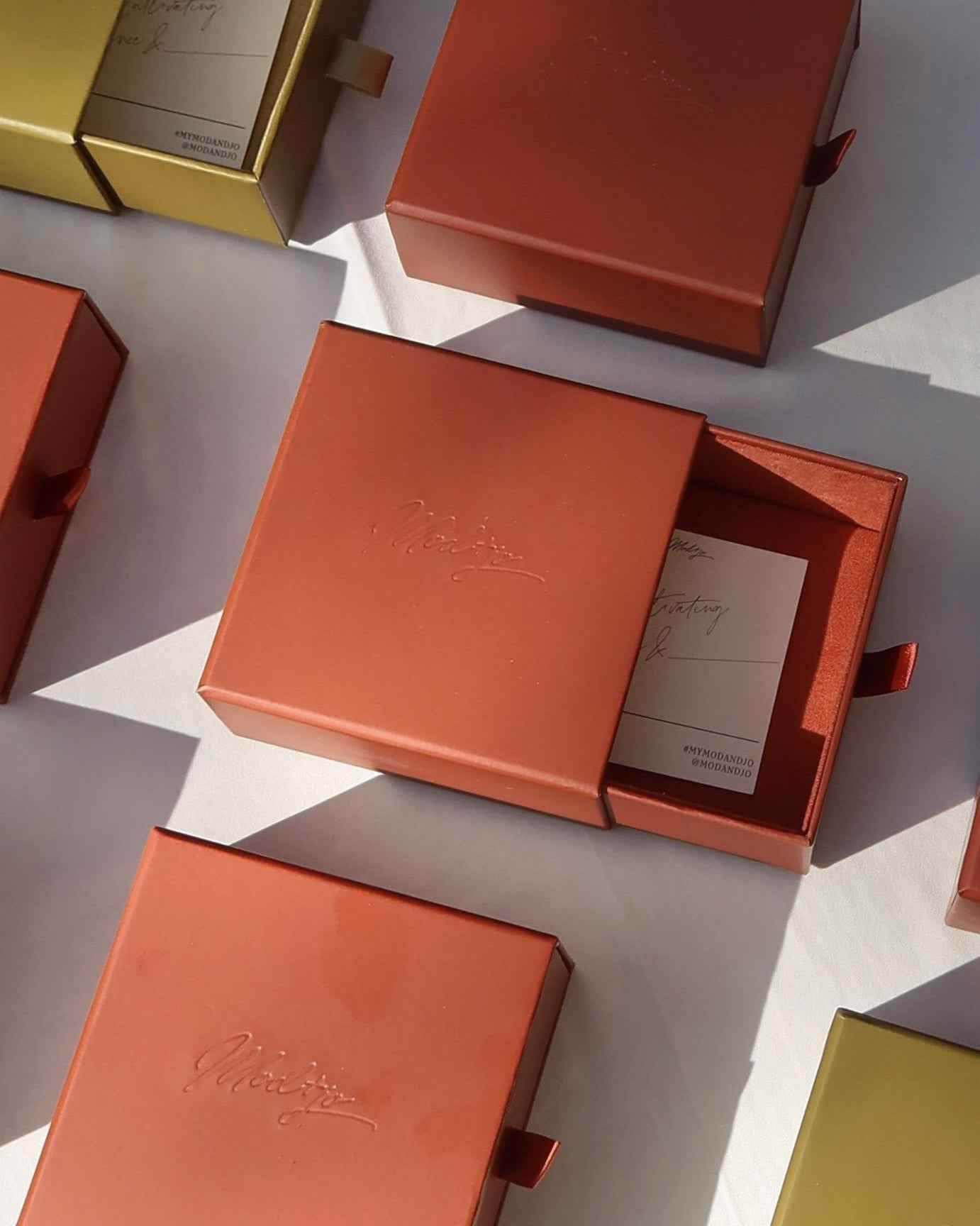 Several red and gold square gift boxes from Mod + Jo sit on a white surface, one open to reveal a card and the Tommy Sapphire Pendant Necklace featuring a lab-grown white sapphire on an adjustable paracord. Soft sunlight casts gentle shadows.