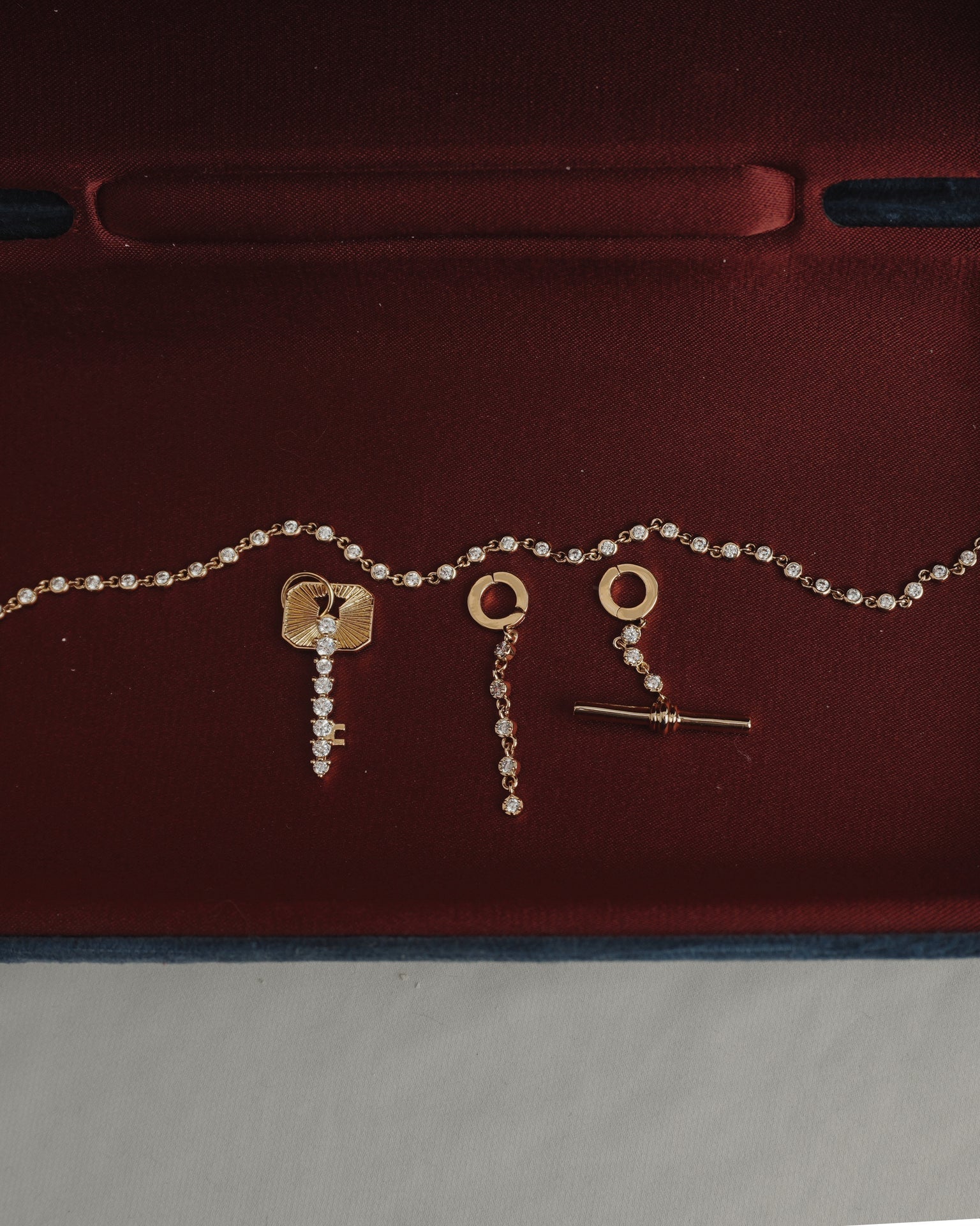 The Mod + Jo Demi Tennis Charm set showcases a dazzling 14k gold-plated chain and matching earrings, both designed with circular tops and delicate dangling accents featuring a sparkling CZ charm, beautifully presented on a deep red velvet backdrop.