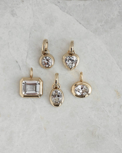 Five Mod + Jo Jovie Sapphire Heart Charms in various gemstone shapes—round, heart, rectangle, oval, and a small round lab-grown white sapphire—are displayed on a light gray marble surface.