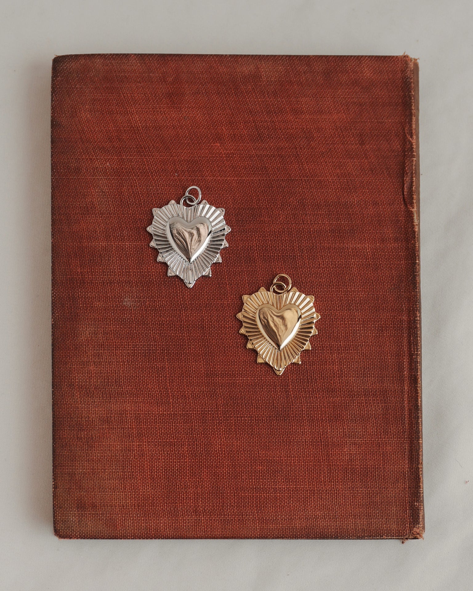 A worn brown book with one silver and one gold heart-shaped medal resting on its cover—both medals featuring a sunburst design behind the heart, reminiscent of Mod + Jo's Grande Lynn Engravable Heart Charm in radiant 14k gold.