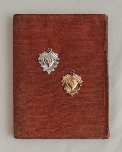 A worn brown book with one silver and one gold heart-shaped medal resting on its cover—both medals featuring a sunburst design behind the heart, reminiscent of Mod + Jo's Grande Lynn Engravable Heart Charm in radiant 14k gold.